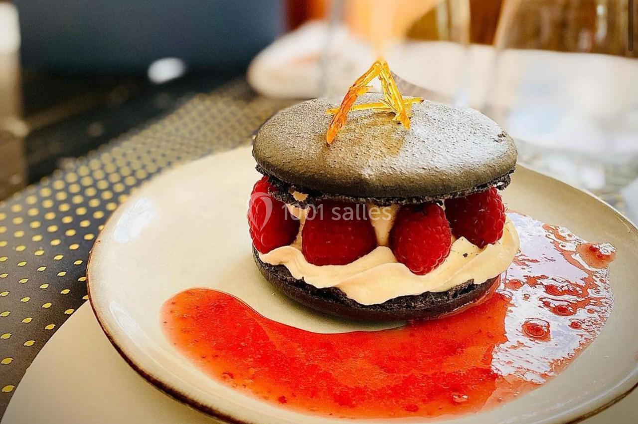 Macaron noir garni de crème et framboises fraîches, accompagné d'un coulis rouge sur une assiette blanche.