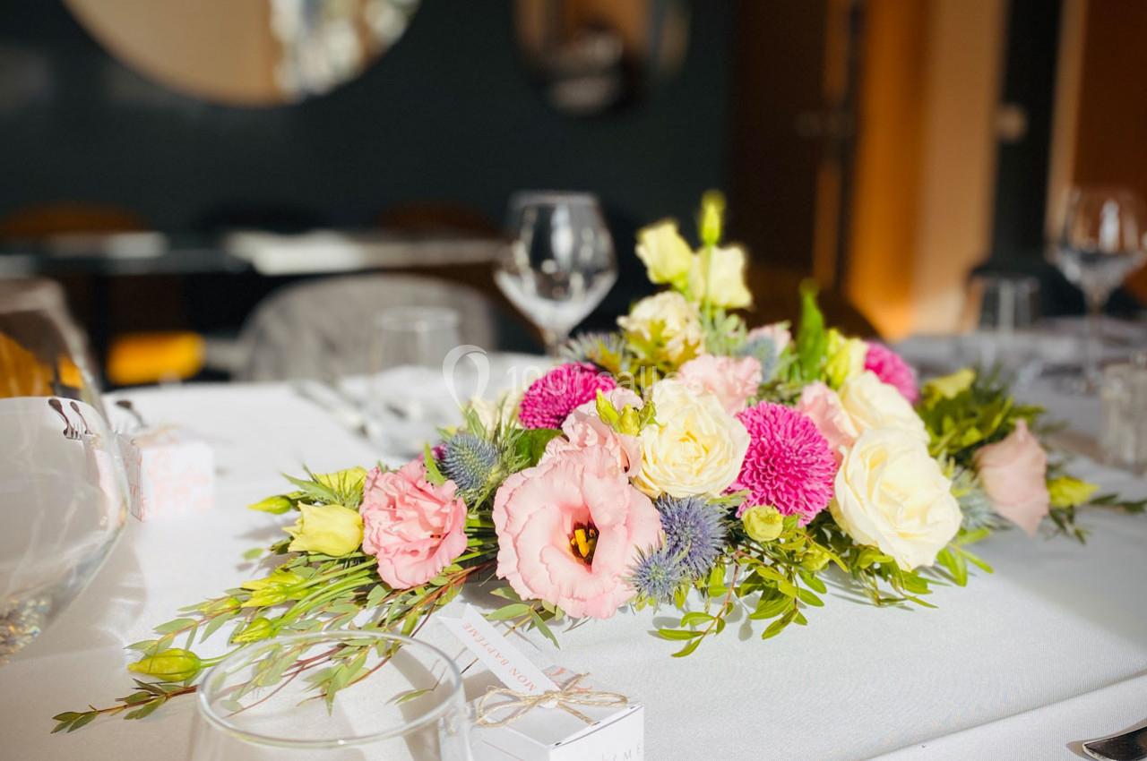 Centre de table floral avec des roses, des chrysanthèmes et des feuillages, posé sur une nappe blanche.
