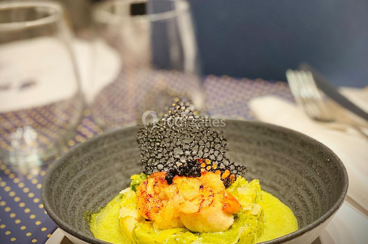 Assiette de crevettes grillées servies sur une purée verte avec une tuile noire décorative, en arrière-plan des verres.