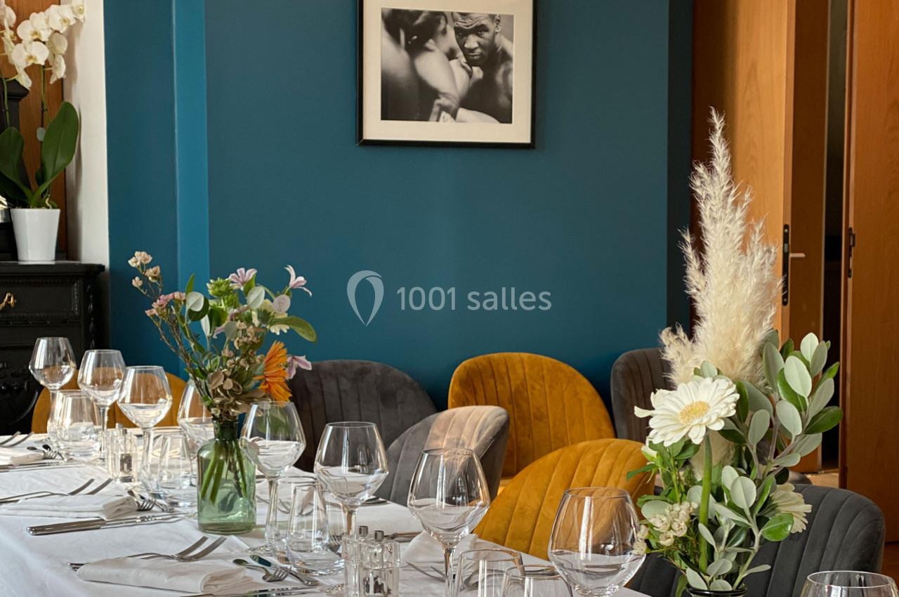 Table dressée avec nappes blanches, verres et fleurs, entourée de chaises colorées dans une salle au mur bleu.