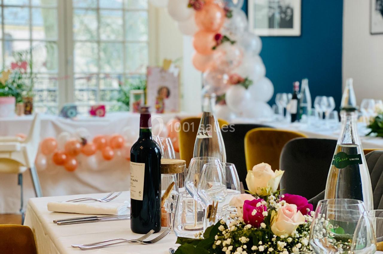 Table dressée pour un événement avec des fleurs, des verres, une bouteille de vin et une décoration de ballons en arrière…