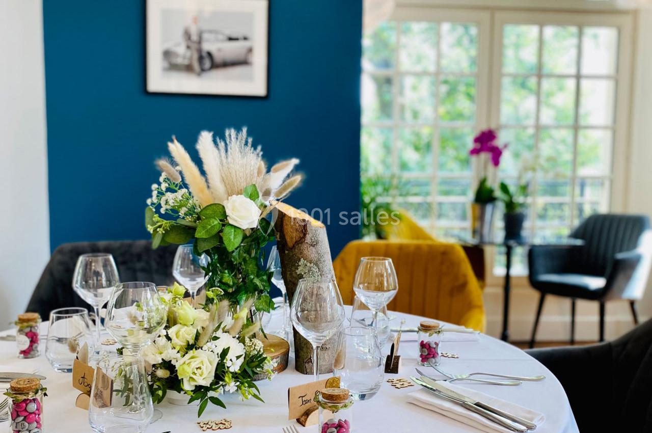 Table élégamment dressée avec des fleurs blanches et pampas, dans une pièce lumineuse aux murs bleus et grandes fenêtres.