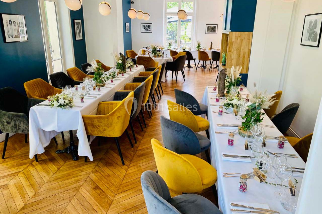 Salle de réception lumineuse avec tables dressées, chaises colorées et décorations florales sur parquet en bois.