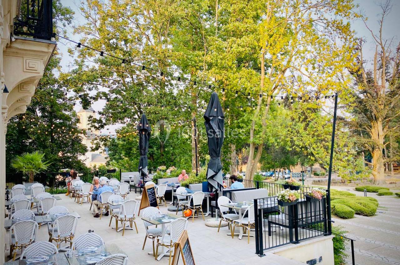 Terrasse d'un café avec des tables et chaises en osier, entourée de verdure et éclairée par des guirlandes lumineuses.