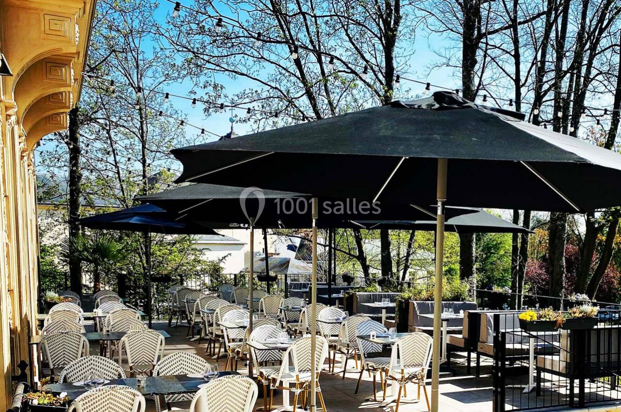 Terrasse ensoleillée avec tables et chaises en osier, ombragée par de grands parasols noirs, entourée d'arbres.