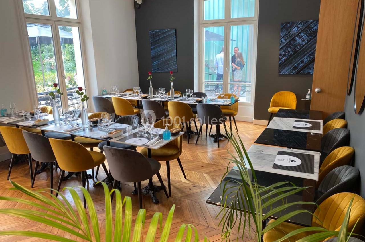 Salle de restaurant lumineuse avec tables dressées, chaises colorées et grandes fenêtres donnant sur un jardin.