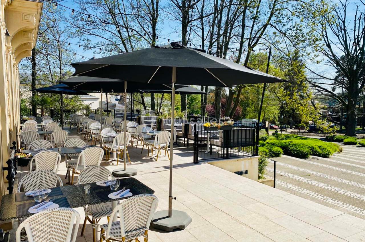 Terrasse d'un restaurant avec tables et chaises en osier, parasols noirs, entourée d'arbres et d'un jardin verdoyant.