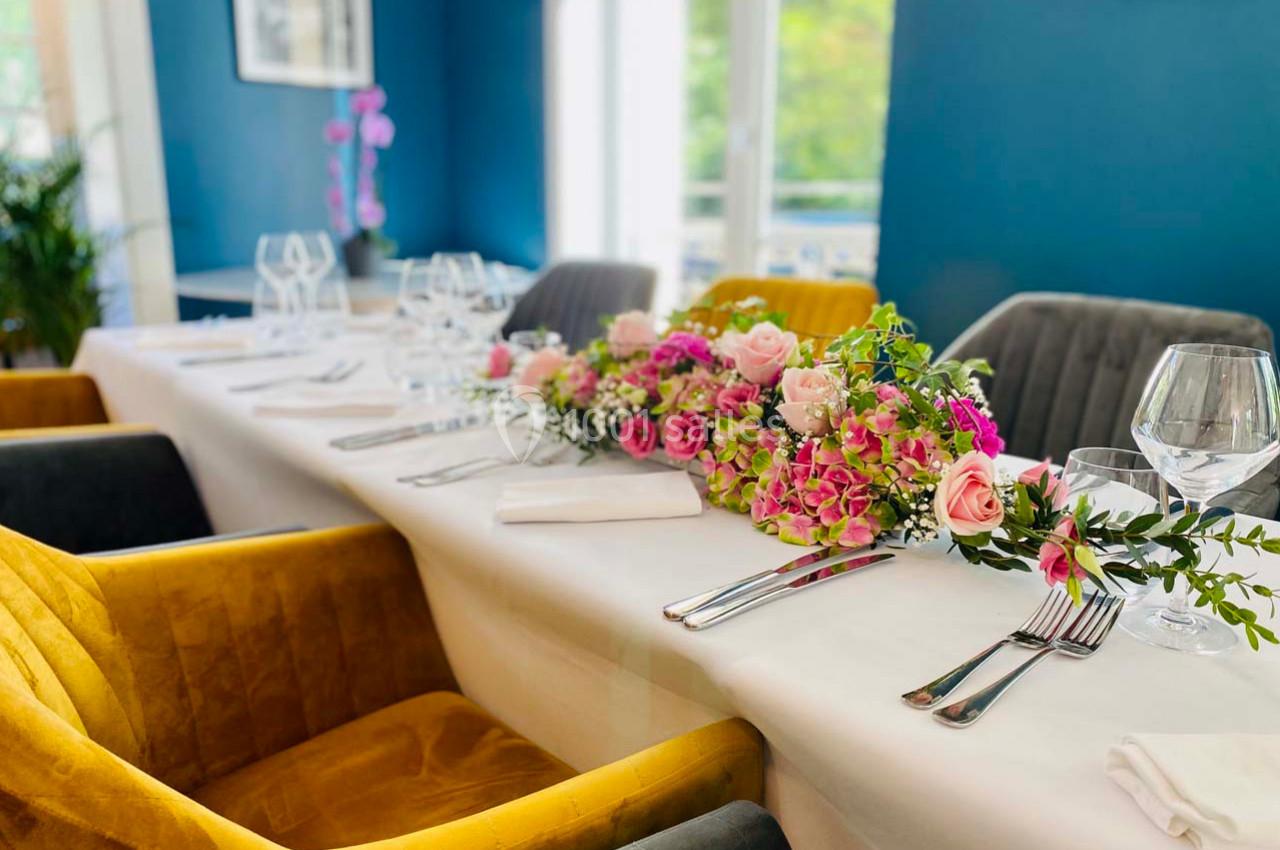 Table élégamment dressée avec nappes blanches, chaises colorées et décoration florale composée de roses et feuillages.