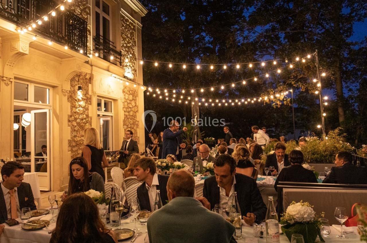 Des convives dînent en soirée sur une terrasse éclairée par des guirlandes lumineuses, près d'un bâtiment élégant.