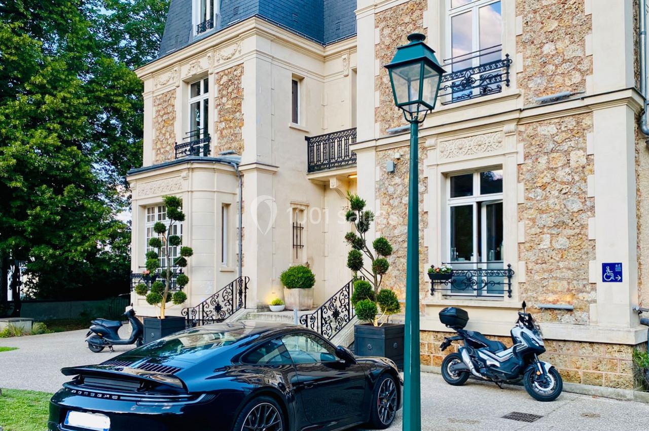 Voiture de sport noire garée devant une maison en pierre avec des escaliers, des plantes et deux scooters à proximité.