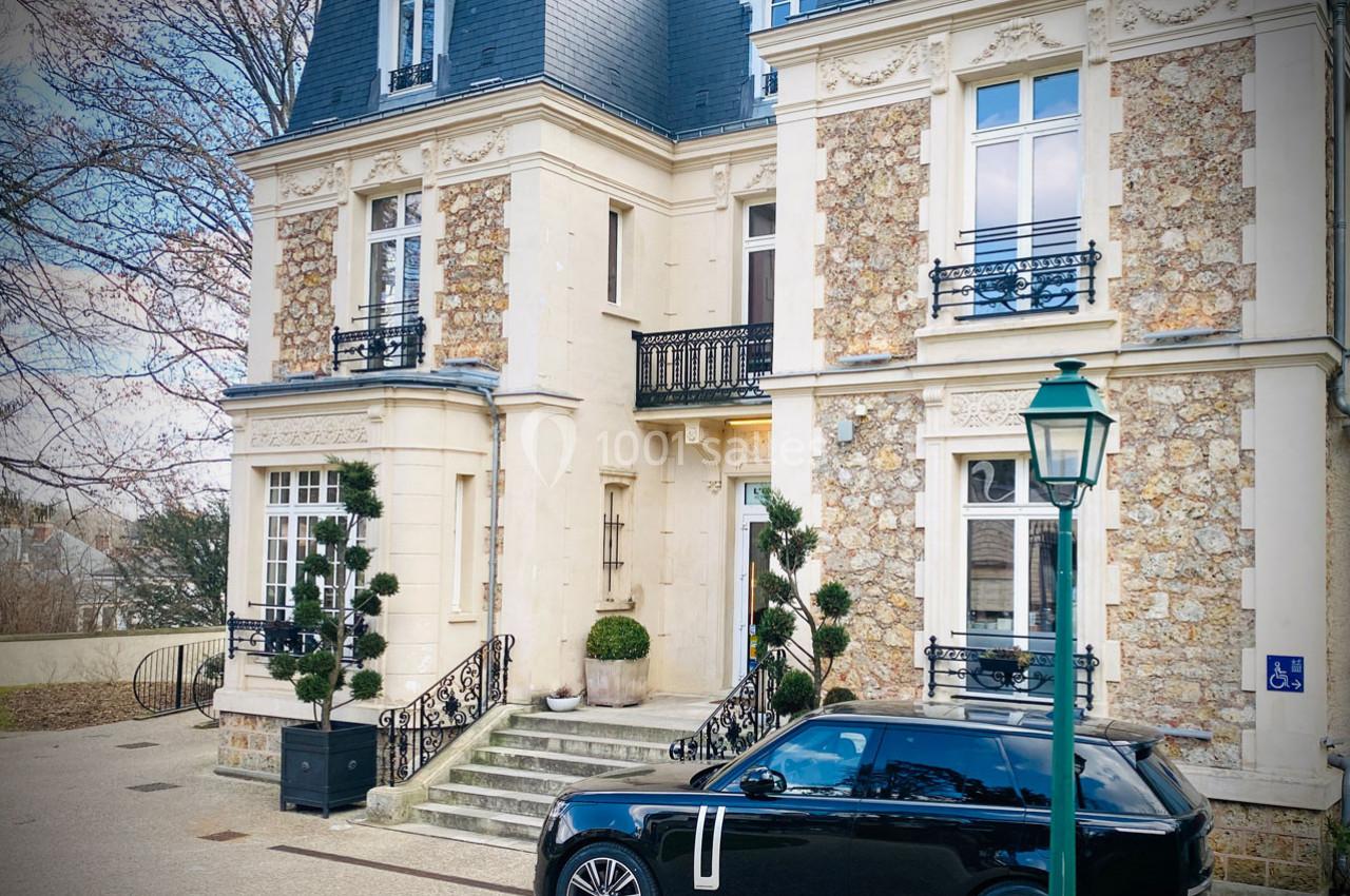 Façade d'une maison bourgeoise en pierre avec un escalier, des plantes taillées et une voiture noire stationnée devant.