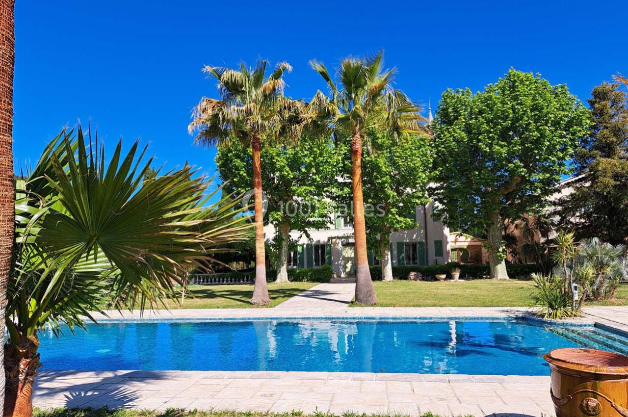 Piscine extérieure entourée de palmiers et d'arbres, avec une maison en arrière-plan sous un ciel bleu clair.