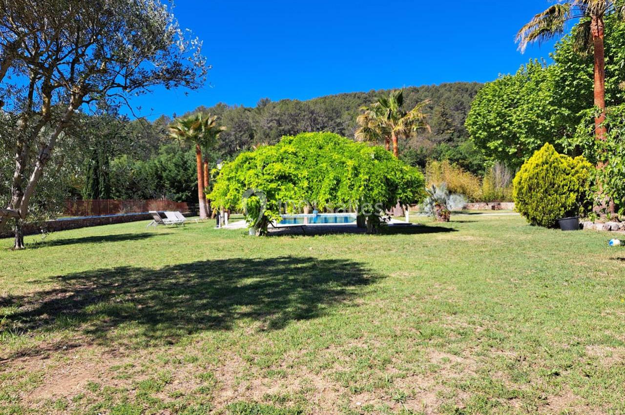 Jardin avec pelouse, arbres, piscine au centre et collines boisées en arrière-plan sous un ciel bleu.