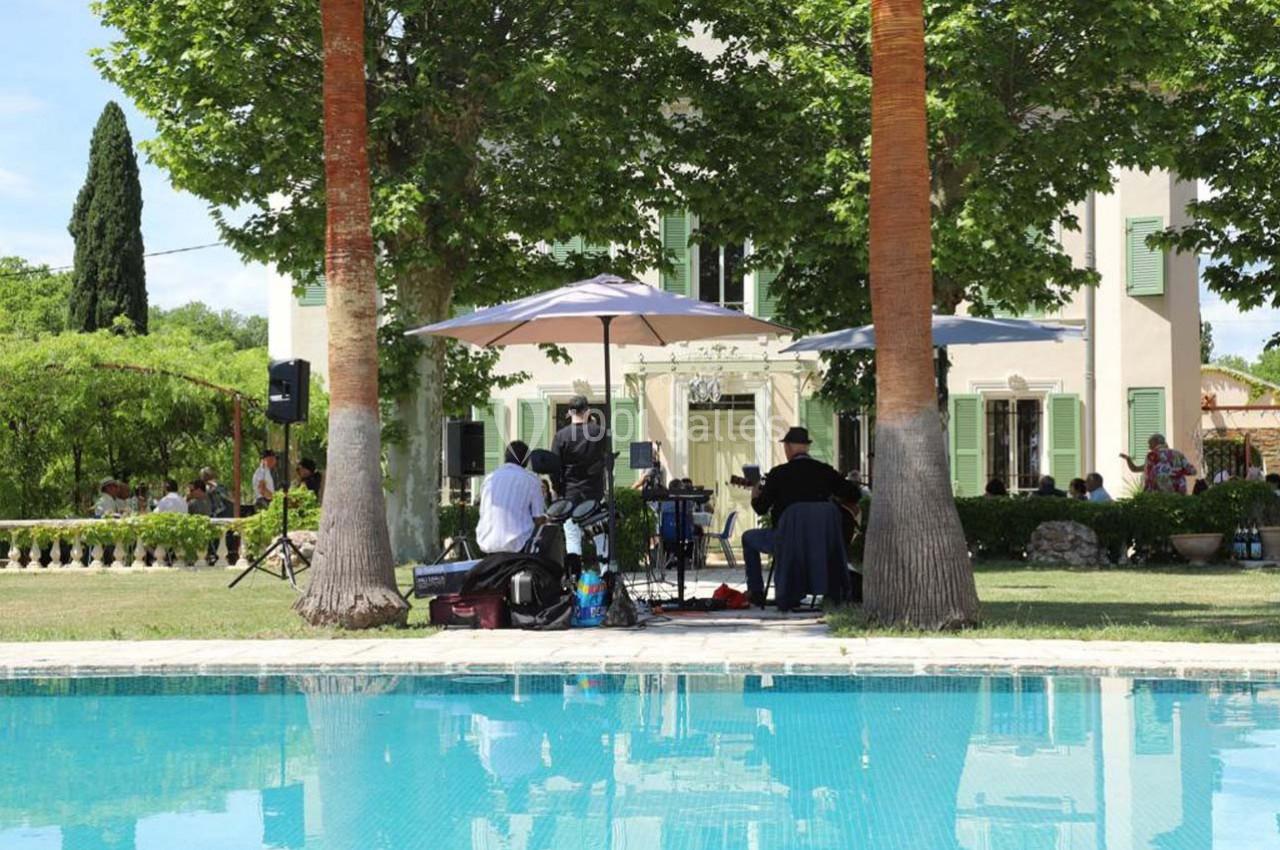 Musiciens jouant sous des parasols dans un jardin, près d'une piscine, devant une maison entourée d'arbres.