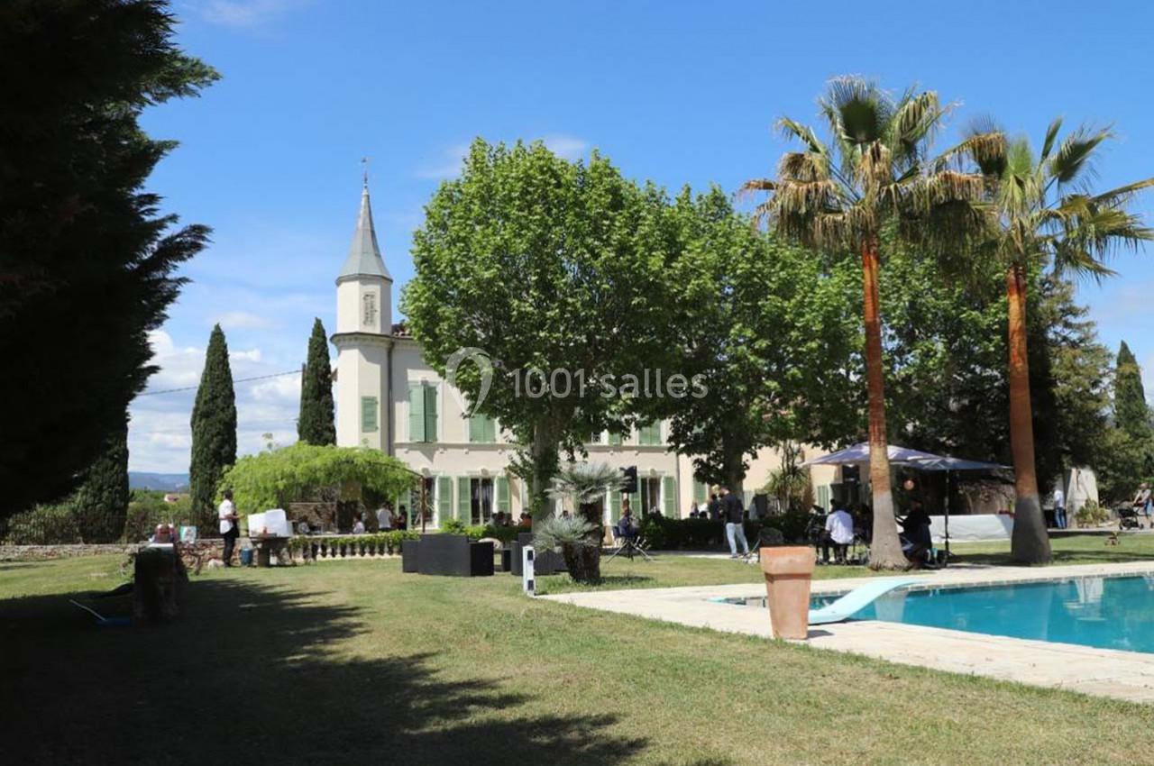 Grande maison avec tour, entourée d'arbres, pelouse et piscine, sous un ciel bleu clair.