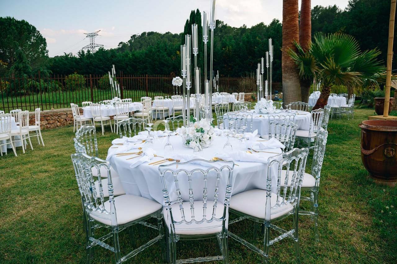 Table ronde élégamment dressée avec nappes blanches, chaises transparentes et centre de table floral, dans un jardin…