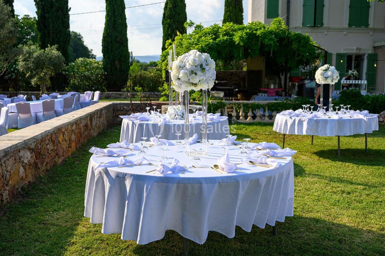 Tables rondes dressées avec nappes blanches et décorations florales dans un jardin verdoyant près d'une maison.
