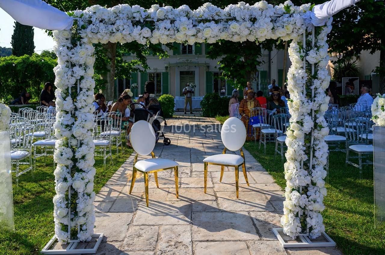Arche de mariage décorée de fleurs blanches, avec deux chaises dorées placées au centre, dans un jardin ensoleillé.