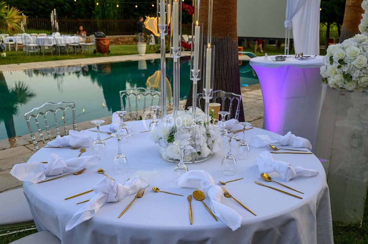 Table élégamment dressée avec des couverts dorés, serviettes blanches et centre de table floral, près d'une piscine.