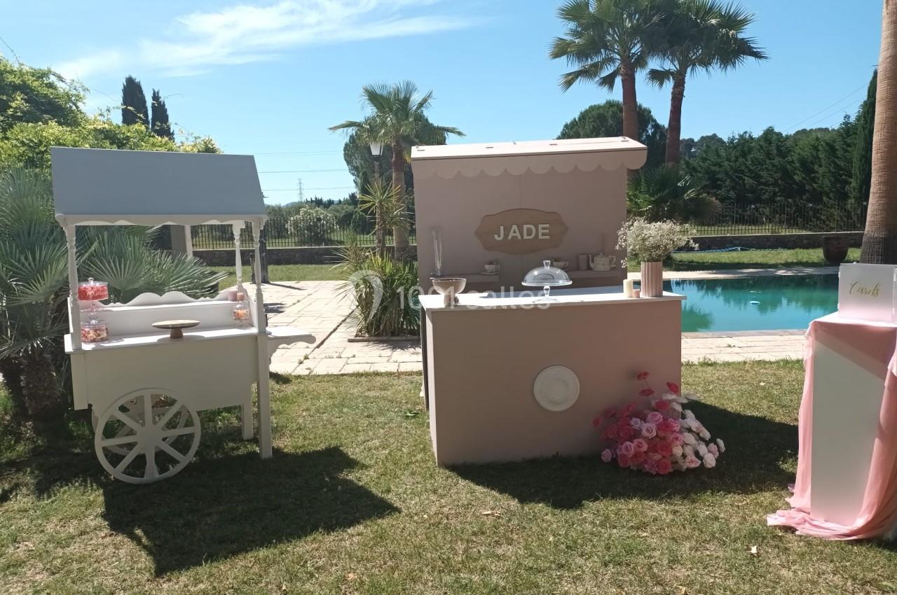 Stand décoratif en extérieur avec chariot blanc, comptoir rose marqué ’Jade’, fleurs et piscine en arrière-plan.