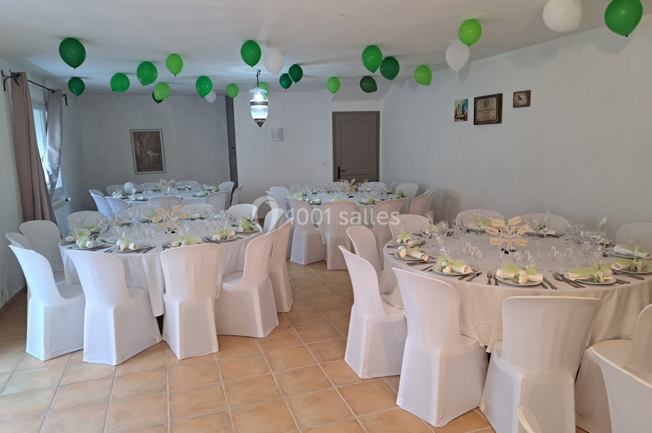 Salle décorée pour un événement avec des tables rondes dressées, nappes blanches et ballons verts suspendus.