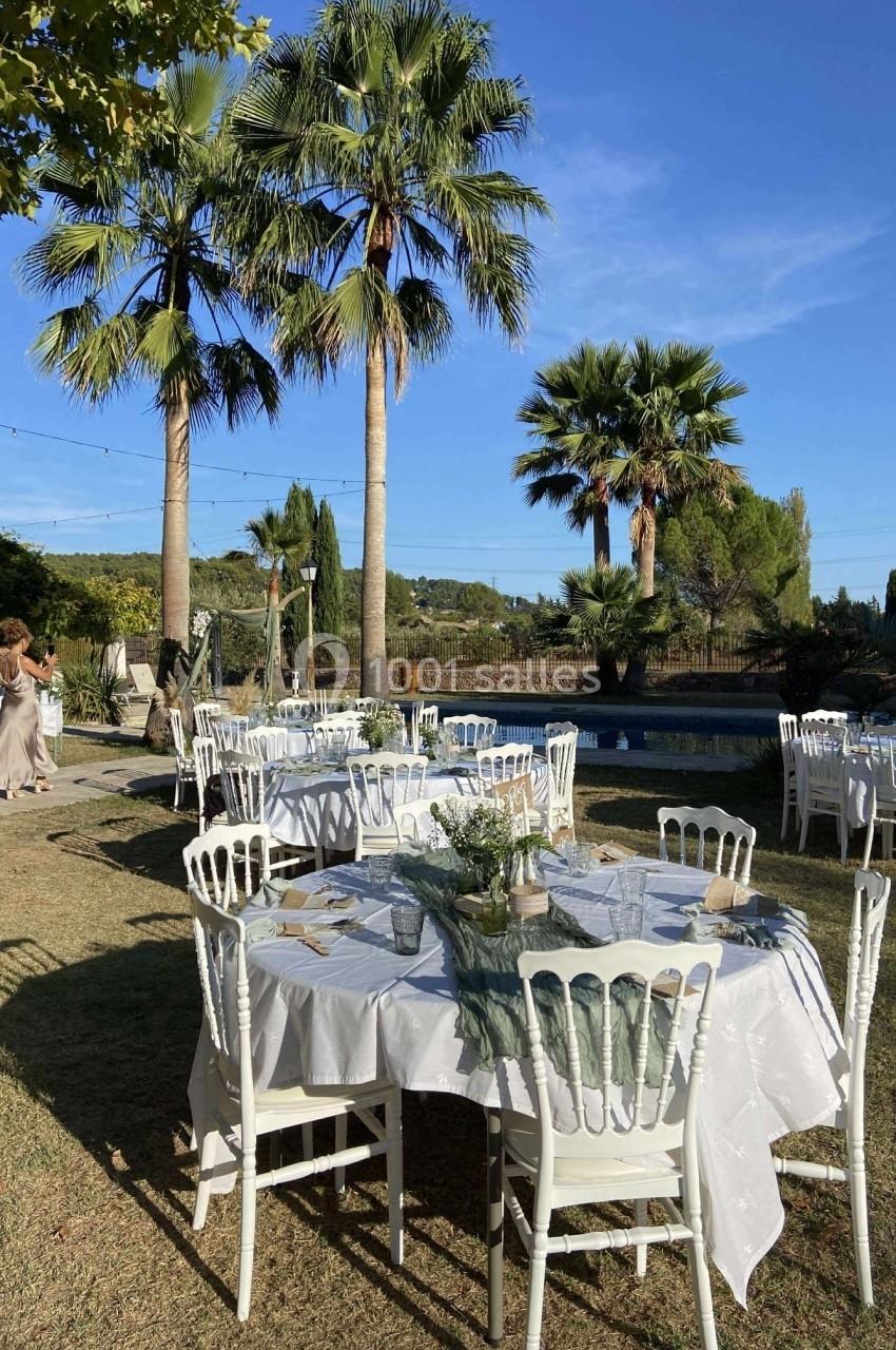 Tables dressées avec nappes blanches et chaises en extérieur, près d'une piscine entourée de palmiers sous un ciel bleu.