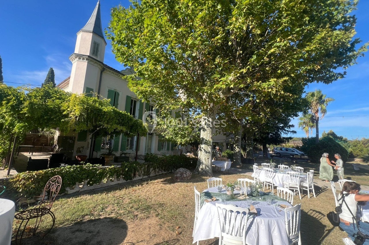 Tables dressées en extérieur sous des arbres, près d'une grande maison avec des volets verts par une journée ensoleillée.