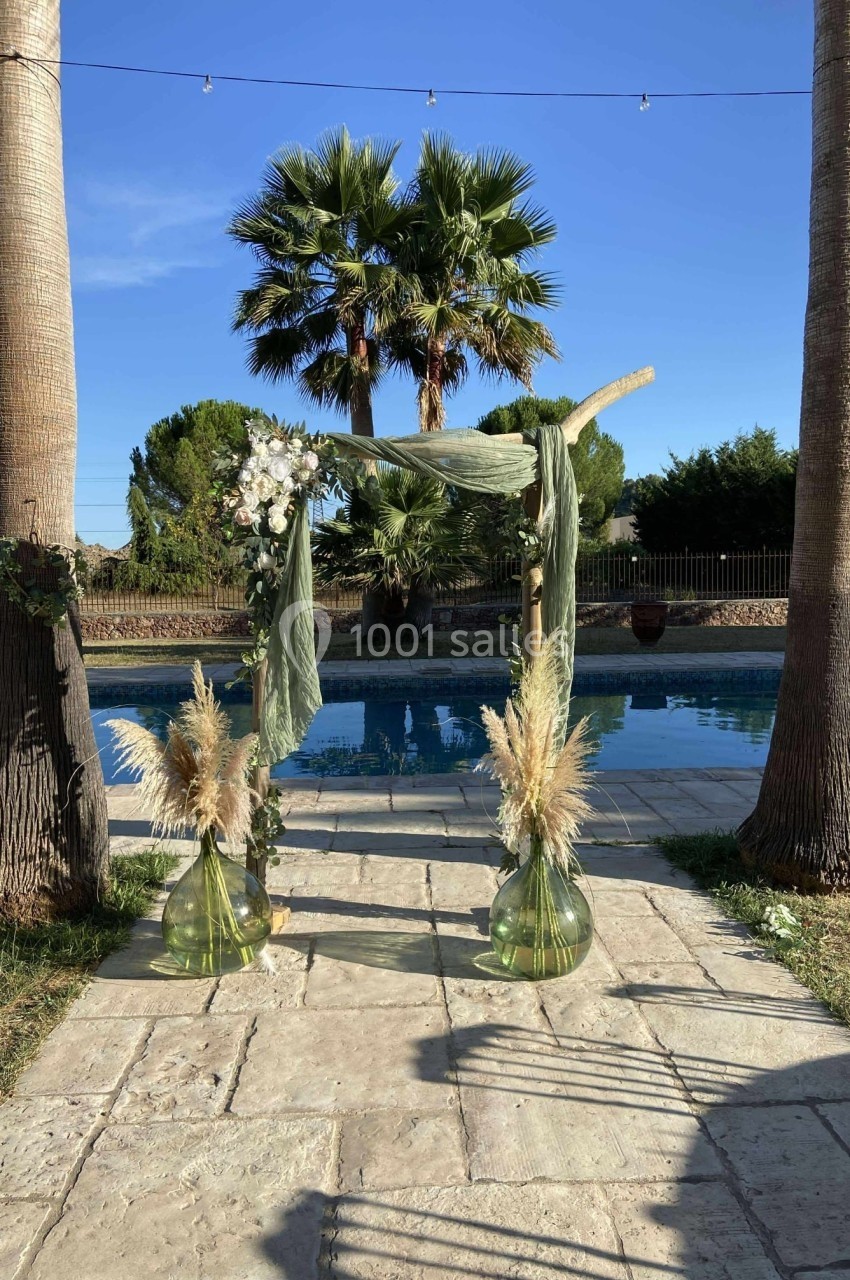 Arche de cérémonie décorée de voilages verts, fleurs et pampas, installée entre deux palmiers près d'une piscine.