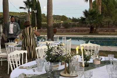 Tables dressées en extérieur avec nappes blanches, entourées de chaises, près d'une piscine et de palmiers sous un ciel bleu.