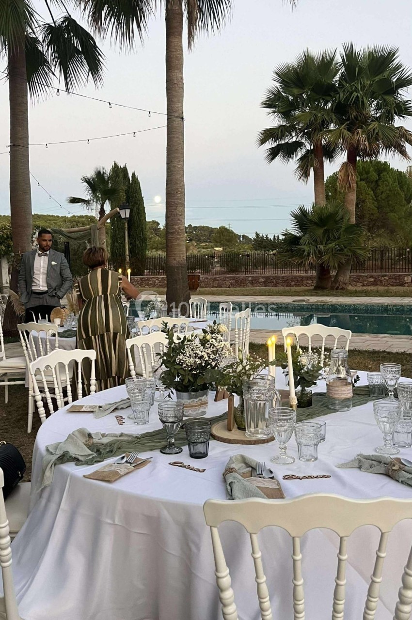 Tables dressées en extérieur avec nappes blanches, décorations florales et bougies, près d'une piscine entourée de palmiers.
