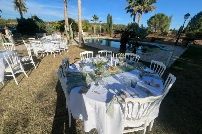 Tables dressées en extérieur avec nappes blanches, entourées de chaises, près d'une piscine et de palmiers sous un ciel bleu.