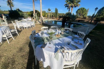 Tables dressées en extérieur avec nappes blanches, entourées de chaises, près d'une piscine et de palmiers sous un ciel bleu.
