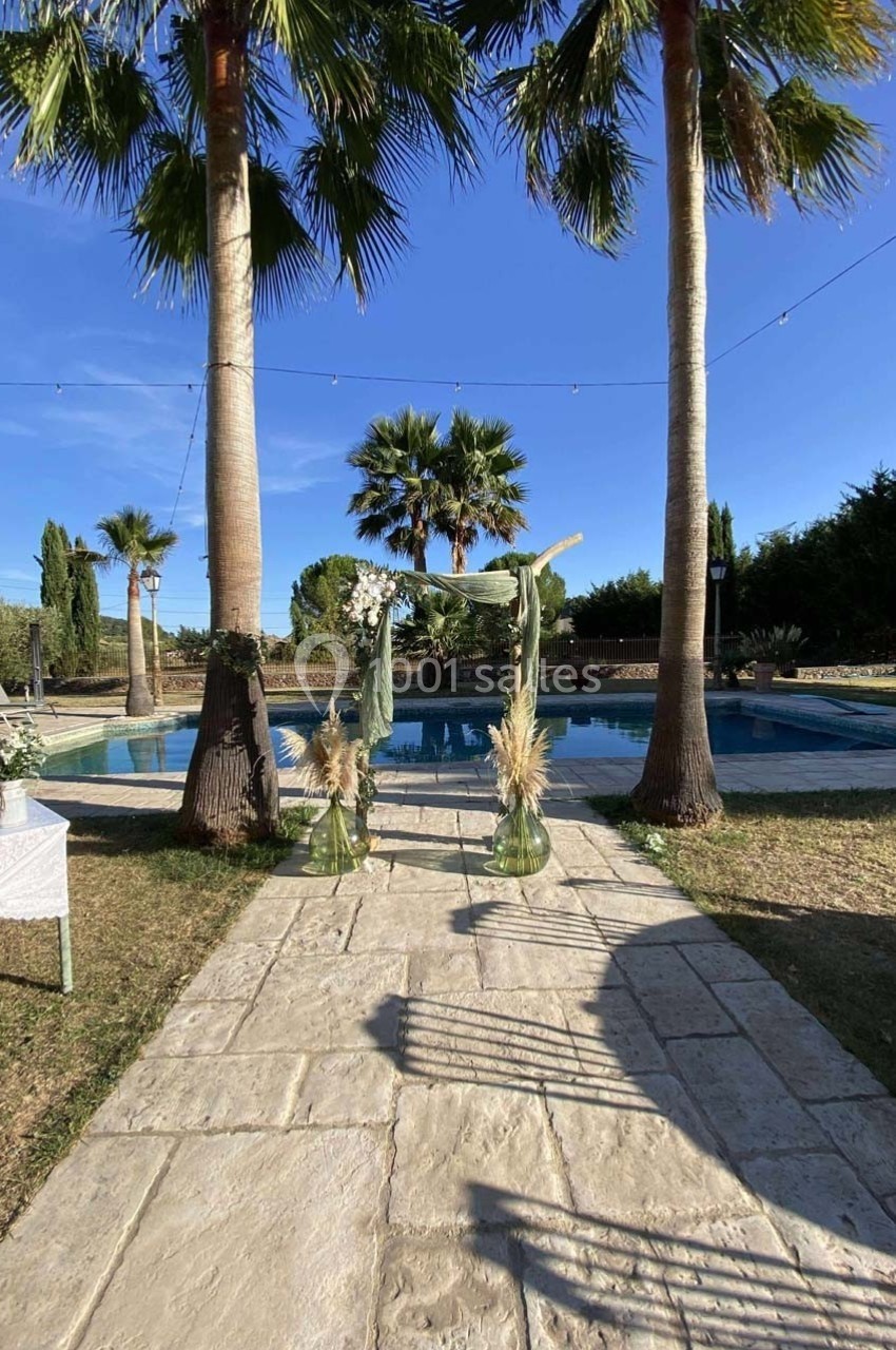 Allée en pierre menant à une arche décorée de feuillages et de fleurs, devant une piscine entourée de palmiers.
