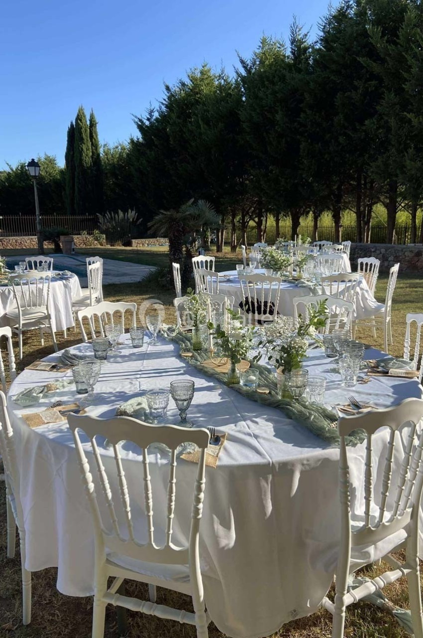 Tables rondes dressées avec nappes blanches, vaisselle et décorations florales dans un jardin bordé d'arbres.