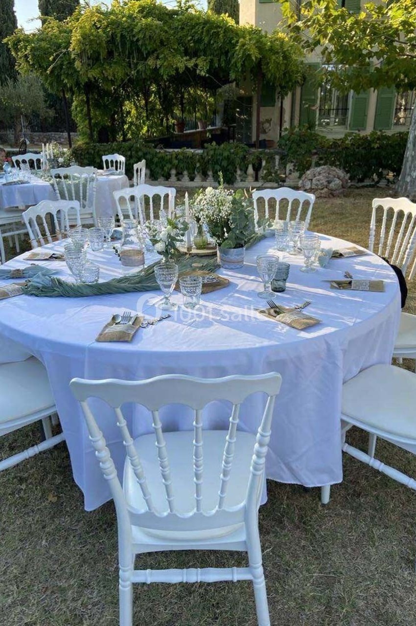 Table ronde dressée en extérieur avec nappe blanche, chaises blanches et décoration florale, sous des arbres.