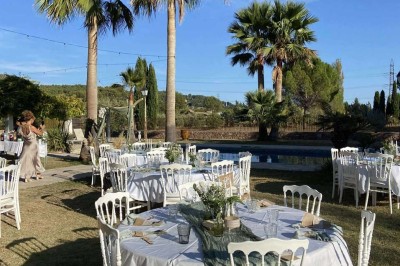 Tables dressées en extérieur avec nappes blanches, entourées de chaises, près d'une piscine et de palmiers sous un ciel bleu.