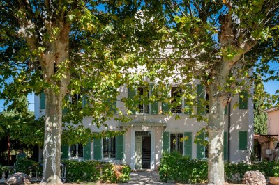 Façade d'une maison provençale blanche avec volets verts, terrasse ombragée et clocher en arrière-plan.