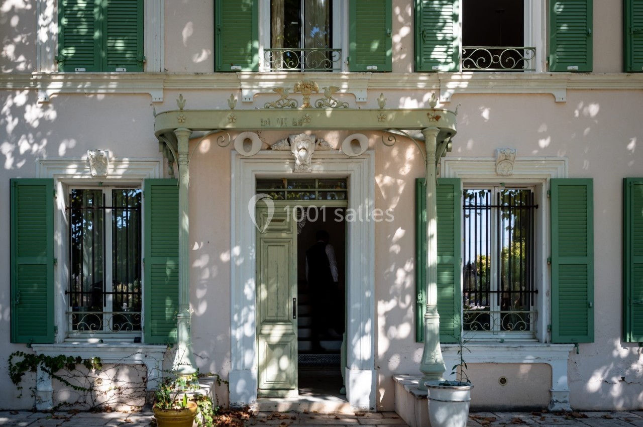 Façade d'une maison claire avec volets verts, porte centrale ornée et ombres d'arbres projetées sur les murs.