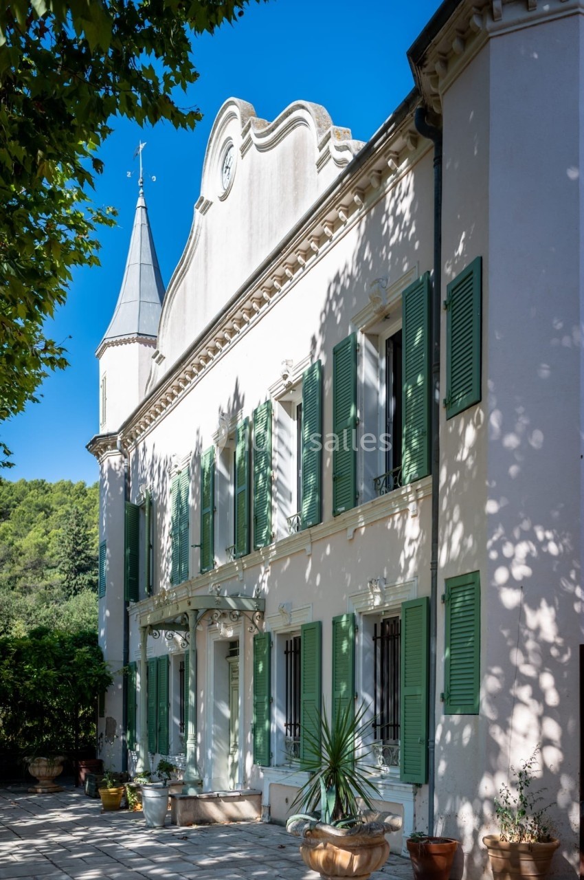 Façade d'une maison provençale blanche avec volets verts, terrasse ombragée et clocher en arrière-plan.