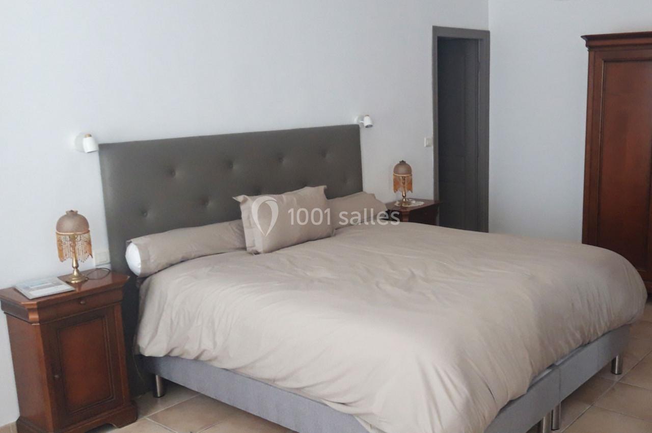 Lit double avec tête de lit grise, draps beiges, flanqué de deux tables de chevet en bois avec lampes.