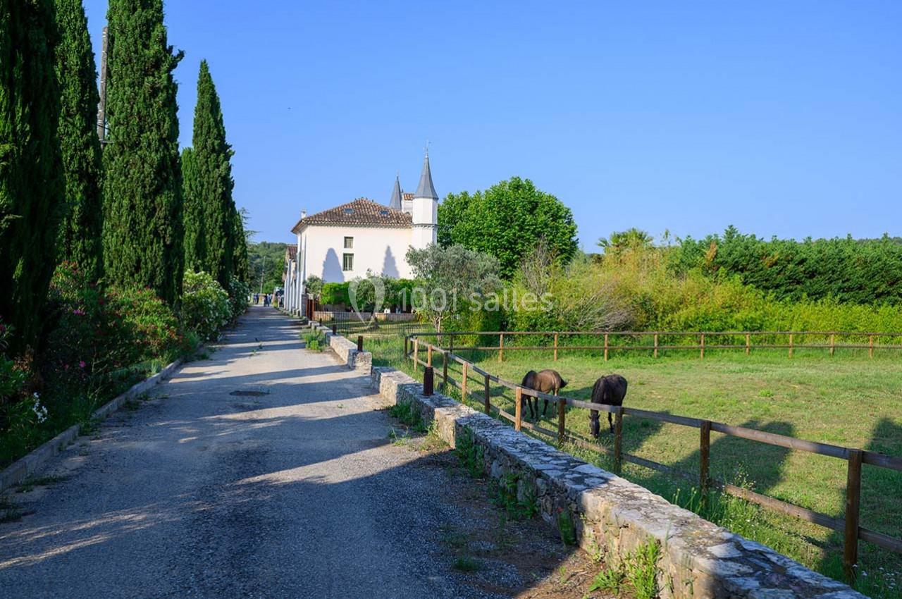 Chemin bordé d'arbres menant à une église blanche, avec un pré où paissent deux chevaux sur la droite.