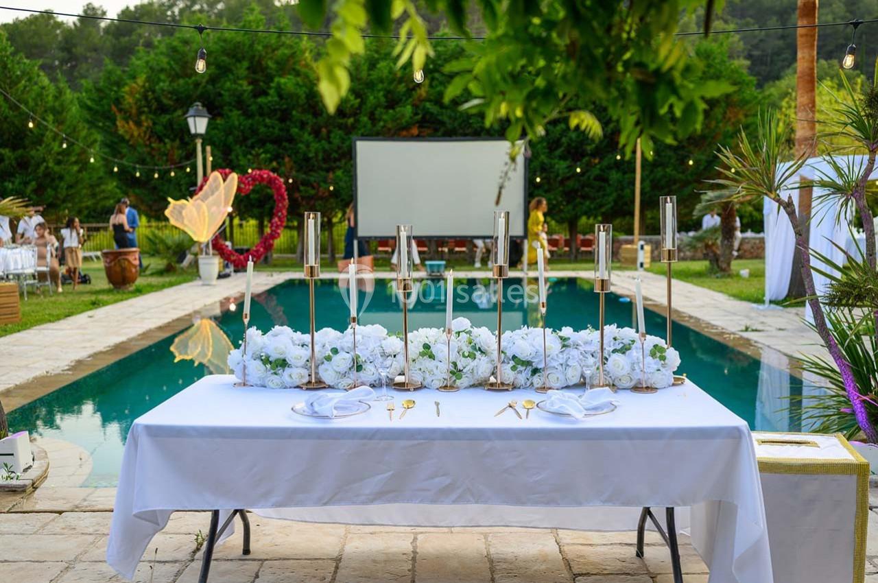 Table décorée avec des fleurs blanches et des bougies, placée devant une piscine entourée de verdure et de guirlandes…