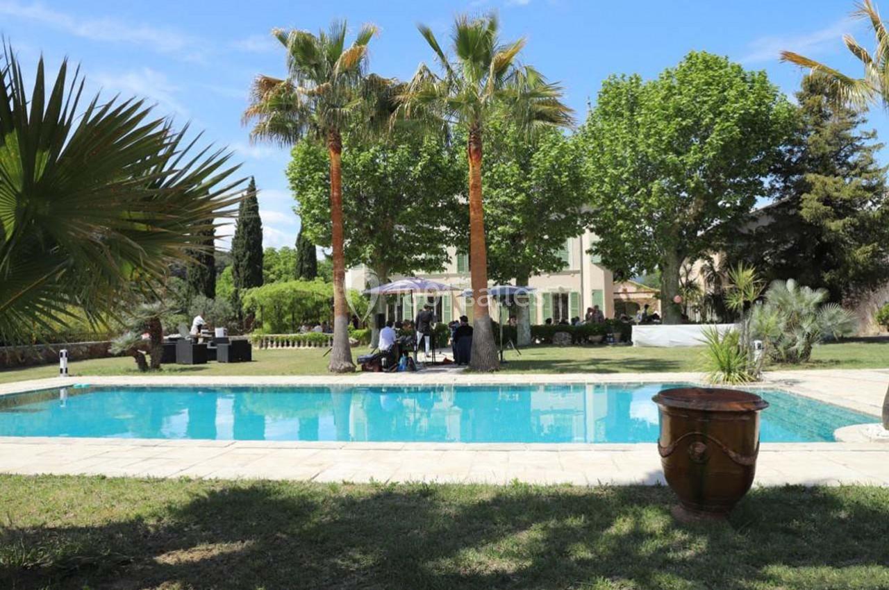 Jardin avec piscine entourée de palmiers et arbres, des personnes assises à des tables sous des parasols.