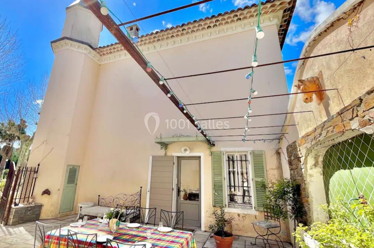 Cour extérieure avec table colorée dressée, chaises en fer forgé, guirlandes lumineuses et façade claire d'une maison.