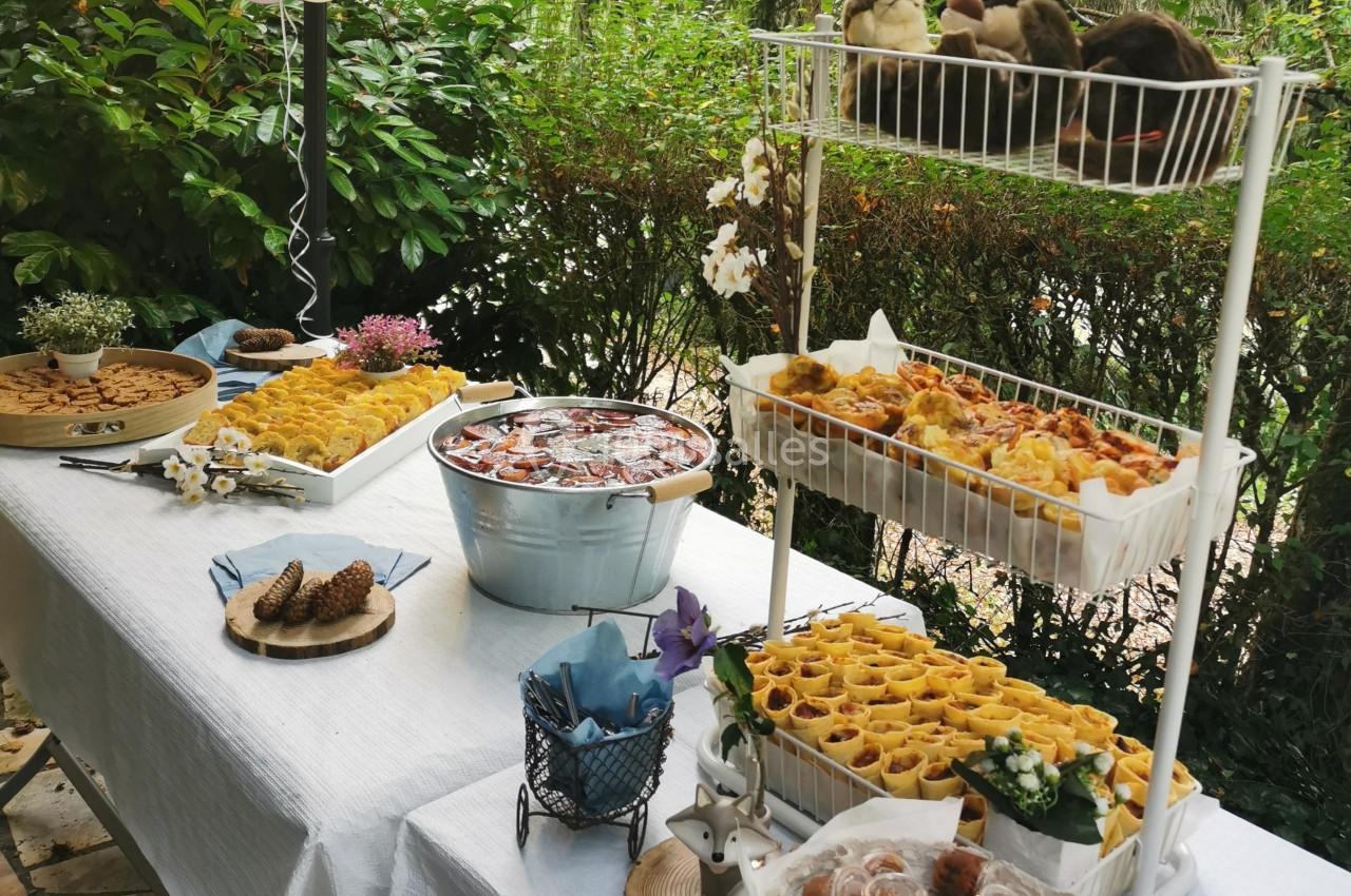 Buffet en extérieur avec plats variés, pâtisseries, boissons dans une bassine et décoration florale sur une nappe blanche.