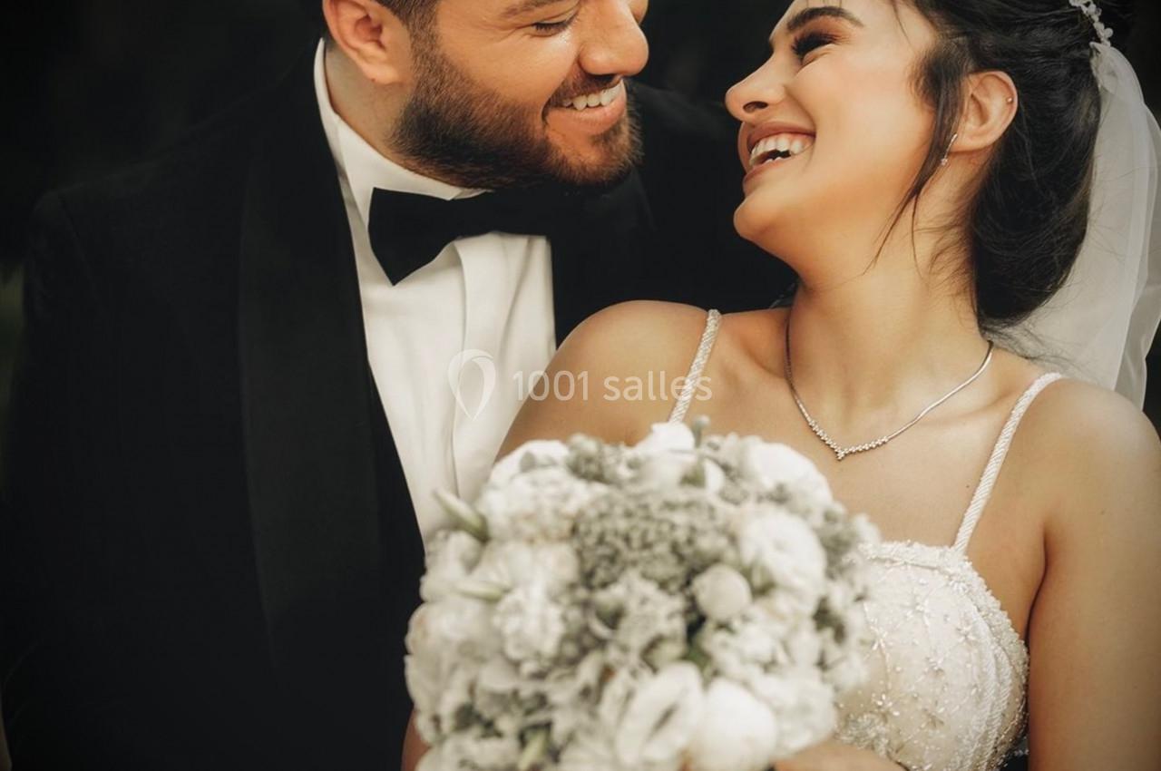 Un couple en tenue de mariage sourit et se regarde tendrement, la mariée tenant un bouquet de fleurs blanches.