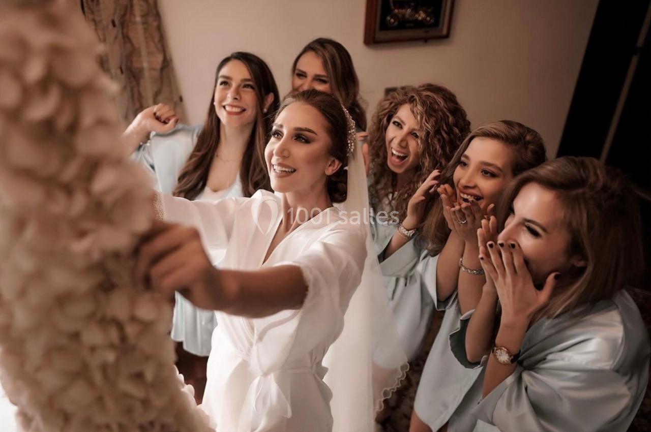 Une mariée en robe blanche entourée de ses demoiselles d'honneur admirant une robe suspendue.