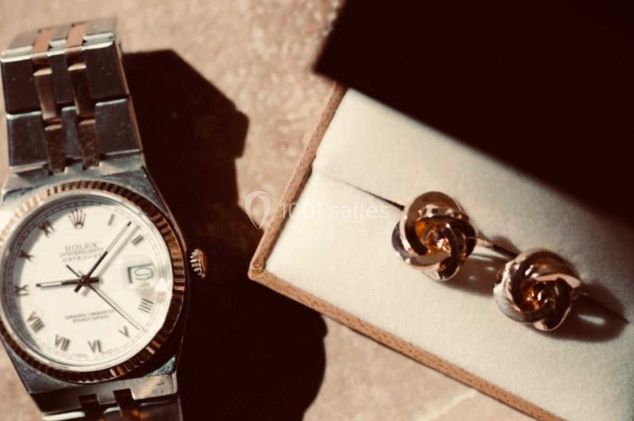 Montre en métal argenté posée sur une surface beige à côté d'une boîte contenant des boutons de manchette dorés.