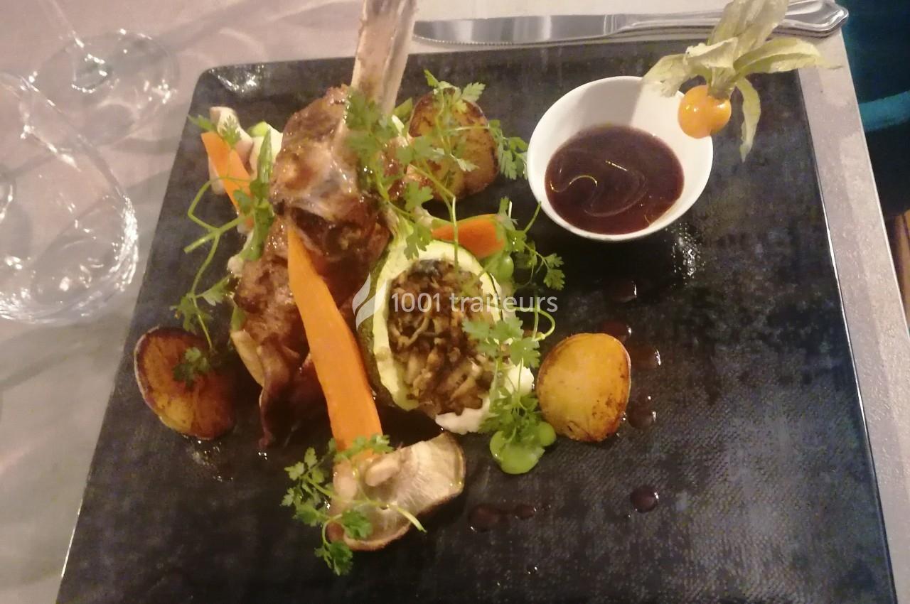 Plat gastronomique composé d'un carré d'agneau, légumes variés, sauce en bol et présentation soignée sur assiette noire.