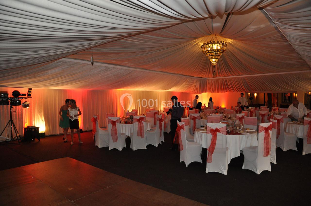 Salle de réception décorée avec des nappes blanches, des chaises ornées de nœuds rouges et un éclairage chaleureux.